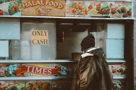 A person stands in front of a food truck with a sign that says 'Only Cash.' The truck advertises various kinds of halal food items such as chicken over rice, lamb over rice, and falafel over rice. The person is wearing a warm jacket and a beanie, suggesting a cold environment, and the truck's images and text are vibrant and colorful.
