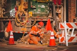 A construction worker wearing an orange safety uniform and helmet is kneeling on the ground, working with tools near a construction vehicle. The area is surrounded by orange and white traffic cones and barriers. Equipment and cables are visible, and the setting appears to be a city street under construction.