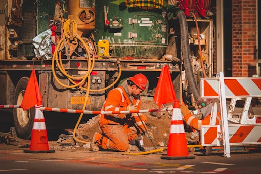 A construction worker wearing an orange safety uniform and helmet is kneeling on the ground, working with tools near a construction vehicle. The area is surrounded by orange and white traffic cones and barriers. Equipment and cables are visible, and the setting appears to be a city street under construction.