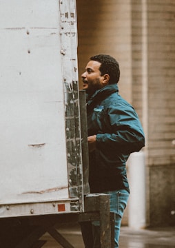 A person standing next to a large white truck, wearing a dark jacket and jeans, appears to be examining the side of the vehicle. The background consists of a brick wall, indicating an urban or industrial setting.