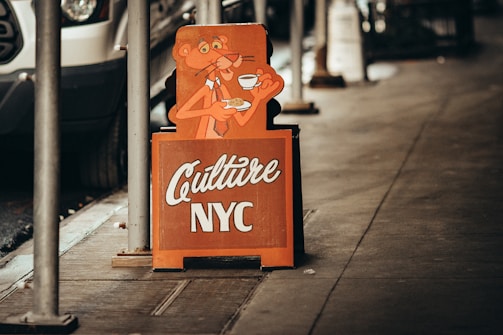 A sidewalk features a sign with an illustration of an orange cartoon character holding a cup and a plate. The sign, with the words 'Culture NYC' in white letters, is positioned near a street with a parked vehicle in partial view.