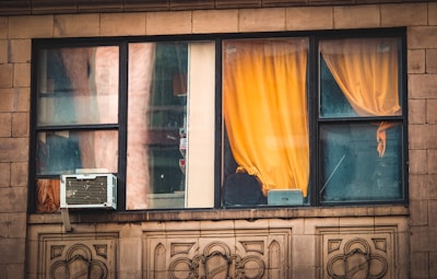 An urban window with a yellow curtain partially drawn, revealing the inside of a building. An air conditioning unit is installed in the lower left segment of the window. The window frame is black and the building exterior is made of stone with decorative patterns. The glass appears slightly weathered.