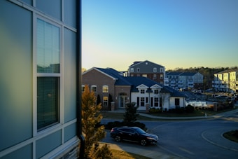 A suburban residential area is depicted with several modern multi-story buildings featuring brick and siding exteriors. The scene is set during the late afternoon with a clear blue sky. Trees and neatly trimmed bushes line the streets, and a black car is parked near the curb.