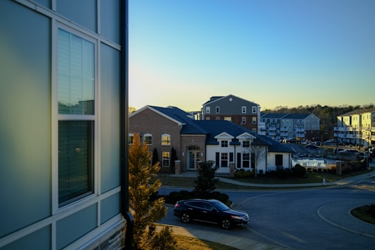 A suburban residential area is depicted with several modern multi-story buildings featuring brick and siding exteriors. The scene is set during the late afternoon with a clear blue sky. Trees and neatly trimmed bushes line the streets, and a black car is parked near the curb.