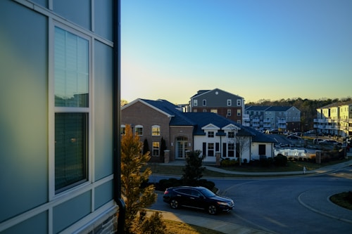 A suburban residential area is depicted with several modern multi-story buildings featuring brick and siding exteriors. The scene is set during the late afternoon with a clear blue sky. Trees and neatly trimmed bushes line the streets, and a black car is parked near the curb.