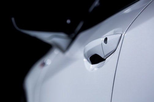 A close-up image of a white car door handle, focusing on its sleek and polished design. The background is dark, emphasizing the glossy surface of the car.