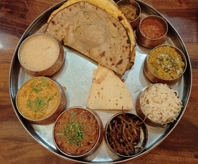A traditional Indian thali is presented on a metal platter containing various small bowls of assorted dishes. There are multiple curries, a serving of rice, a flatbread like roti or naan, a piece of papadum, and possibly a sweet or pickle.