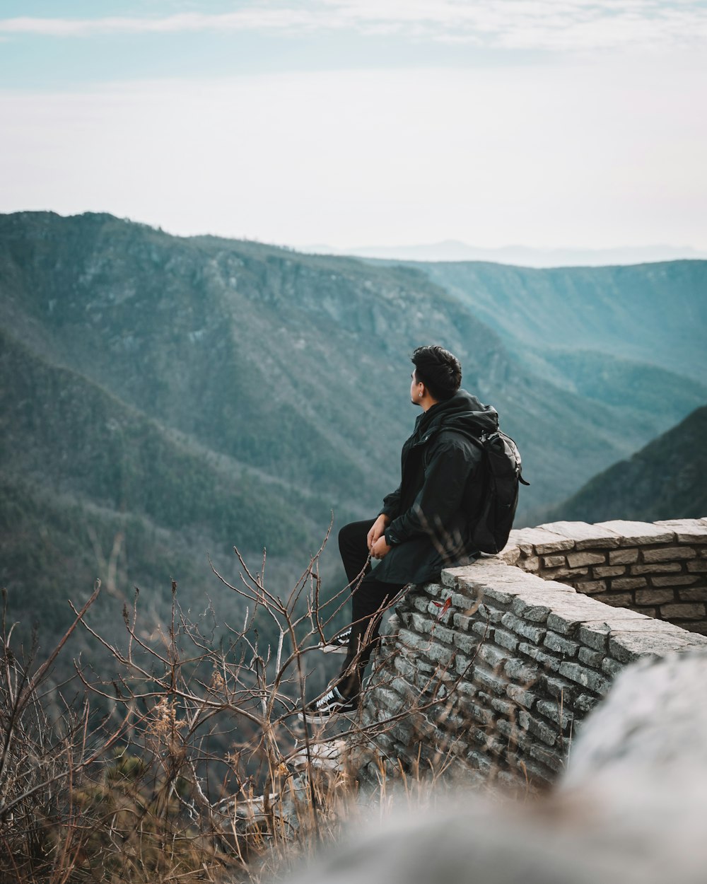 man in black jacket and black backpack sitting on gray concrete wall during daytime