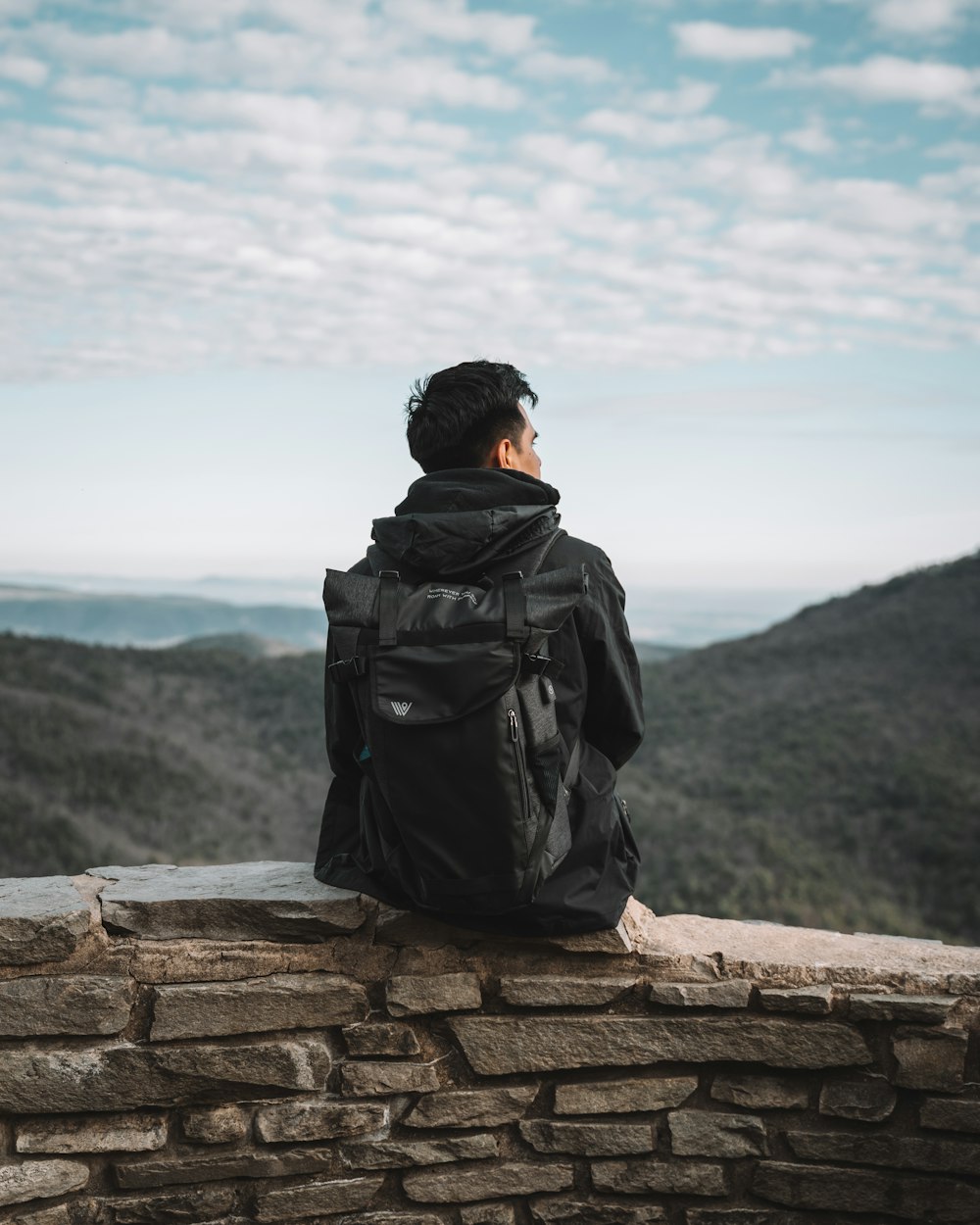 man in black jacket and black backpack sitting on brown concrete wall during daytime