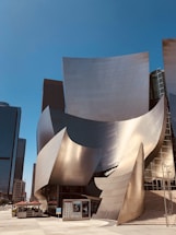 A modern building with a unique, curvilinear design featuring shiny, metallic surfaces. The structure is surrounded by skyscrapers, reflecting sunlight in various directions. There is a small outdoor dining area with umbrellas and a set of steps leading to the entrance.