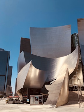 A modern building with a unique, curvilinear design featuring shiny, metallic surfaces. The structure is surrounded by skyscrapers, reflecting sunlight in various directions. There is a small outdoor dining area with umbrellas and a set of steps leading to the entrance.