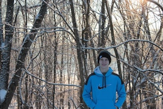 A person in a blue hoodie with a Nike logo and a black beanie stands amidst a forest of tall trees. The branches are lightly coated in snow, and sunlight filters through, casting a warm glow.
