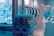 A store display features a mannequin wearing a bunny mask and a turquoise T-shirt, seated near a teal vending machine. The background includes hanging shirts in various colors, mostly white and turquoise. Neon blue signage is on the wall, contributing to a vibrant, modern atmosphere.