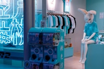 A store display features a mannequin wearing a bunny mask and a turquoise T-shirt, seated near a teal vending machine. The background includes hanging shirts in various colors, mostly white and turquoise. Neon blue signage is on the wall, contributing to a vibrant, modern atmosphere.
