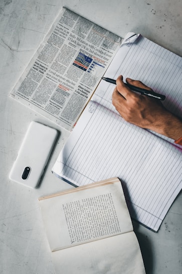 A person holding a pen writes in a lined notebook on a gray surface. A newspaper is spread out to the left, featuring headlines about Covid-19. Below the notebook, a smartphone with a white case is placed, and an open book lies towards the bottom of the image, showing a page of printed text.