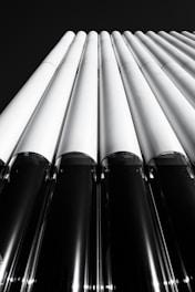 A monochromatic architectural image featuring a series of vertical, cylindrical structures that appear to be part of a building facade. The perspective is from below, looking up towards the sky, emphasizing the height and linearity of the design.