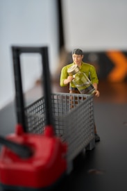 A toy figure of a person dressed in a green shirt holding a container, positioned next to a small, red-handled cart or machine. The setting is indoors, with a background that appears to be a blurred room with bright lighting and a partially visible sign with an arrow.