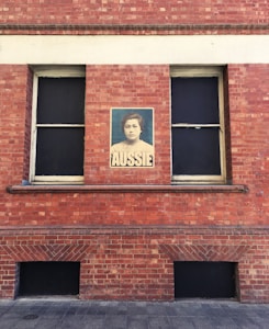 A red brick wall with two windows, one of which has a vintage-style monochromatic poster of a person with 'AUSSIE' written beneath it.