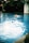 A person is swimming in a blue pool, creating splashes and ripples on the water's surface. Another individual is in the background, appearing to watch or assist. The environment around the pool is lush with greenery, and there is a white structure in the corner.