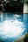 A person is swimming in a blue pool, creating splashes and ripples on the water's surface. Another individual is in the background, appearing to watch or assist. The environment around the pool is lush with greenery, and there is a white structure in the corner.