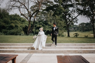 A couple in formal attire is pictured outdoors, with the woman wearing a white wedding dress and holding a bouquet, while the man is dressed in a black suit. They are positioned on white steps amidst a lush green park landscape with trees in the background.