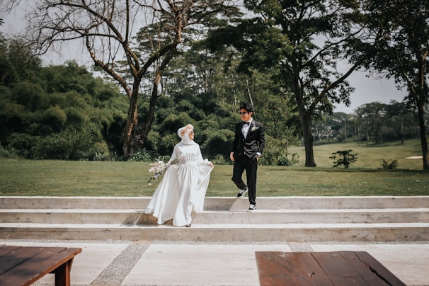 A couple in formal attire is pictured outdoors, with the woman wearing a white wedding dress and holding a bouquet, while the man is dressed in a black suit. They are positioned on white steps amidst a lush green park landscape with trees in the background.