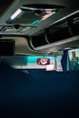 The image captures the interior of a vehicle, likely a bus or coach, showing the ceiling and overhead compartments. A driver's reflection is visible in a rearview mirror wearing a face mask. There are minimal objects and the area is dimly lit, with some natural light filtering through the windows.