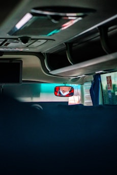The image captures the interior of a vehicle, likely a bus or coach, showing the ceiling and overhead compartments. A driver's reflection is visible in a rearview mirror wearing a face mask. There are minimal objects and the area is dimly lit, with some natural light filtering through the windows.