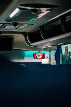 The image captures the interior of a vehicle, likely a bus or coach, showing the ceiling and overhead compartments. A driver's reflection is visible in a rearview mirror wearing a face mask. There are minimal objects and the area is dimly lit, with some natural light filtering through the windows.