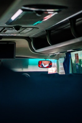 The image captures the interior of a vehicle, likely a bus or coach, showing the ceiling and overhead compartments. A driver's reflection is visible in a rearview mirror wearing a face mask. There are minimal objects and the area is dimly lit, with some natural light filtering through the windows.