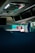 The image captures the interior of a vehicle, likely a bus or coach, showing the ceiling and overhead compartments. A driver's reflection is visible in a rearview mirror wearing a face mask. There are minimal objects and the area is dimly lit, with some natural light filtering through the windows.