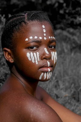A young person with face paint in white patterns is looking directly at the camera. The background is blurred with natural surroundings, highlighting the individual's expression and detailed face paint designs.