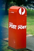 A red Australia Post mailbox stands on a sidewalk surrounded by grass and a few trees in the background. The mailbox features the Australia Post logo in white, which prominently contrasts with its red color.