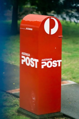 A red Australia Post mailbox stands on a sidewalk surrounded by grass and a few trees in the background. The mailbox features the Australia Post logo in white, which prominently contrasts with its red color.