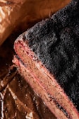 A close-up of a cooked meat slice with a charred and seasoned crust. The meat appears juicy and tender, with a visible smoke ring near the edges. It rests on brown parchment paper, likely part of a barbecue dish.