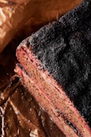 A close-up of a cooked meat slice with a charred and seasoned crust. The meat appears juicy and tender, with a visible smoke ring near the edges. It rests on brown parchment paper, likely part of a barbecue dish.