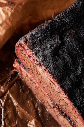 A close-up of a cooked meat slice with a charred and seasoned crust. The meat appears juicy and tender, with a visible smoke ring near the edges. It rests on brown parchment paper, likely part of a barbecue dish.
