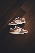 A pair of brown and white sneakers with prominent branding positioned on a dark wooden floor. The stylish shoes have a low-top design with white shoelaces and a distinctive swoosh logo on the sides.