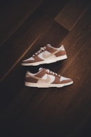 A pair of brown and white sneakers with prominent branding positioned on a dark wooden floor. The stylish shoes have a low-top design with white shoelaces and a distinctive swoosh logo on the sides.