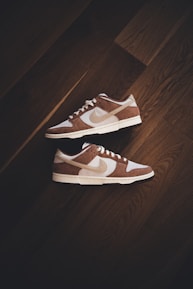 A pair of brown and white sneakers with prominent branding positioned on a dark wooden floor. The stylish shoes have a low-top design with white shoelaces and a distinctive swoosh logo on the sides.