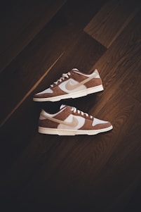 A pair of brown and white sneakers with prominent branding positioned on a dark wooden floor. The stylish shoes have a low-top design with white shoelaces and a distinctive swoosh logo on the sides.