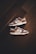 A pair of brown and white sneakers with prominent branding positioned on a dark wooden floor. The stylish shoes have a low-top design with white shoelaces and a distinctive swoosh logo on the sides.