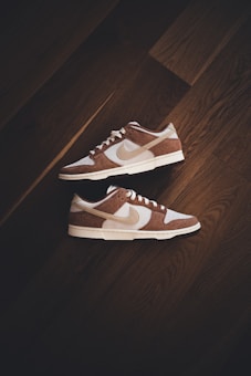 A pair of brown and white sneakers with prominent branding positioned on a dark wooden floor. The stylish shoes have a low-top design with white shoelaces and a distinctive swoosh logo on the sides.