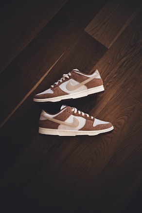 A pair of brown and white sneakers with prominent branding positioned on a dark wooden floor. The stylish shoes have a low-top design with white shoelaces and a distinctive swoosh logo on the sides.