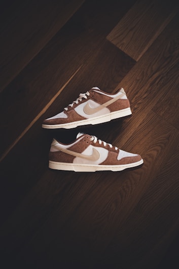 A pair of brown and white sneakers with prominent branding positioned on a dark wooden floor. The stylish shoes have a low-top design with white shoelaces and a distinctive swoosh logo on the sides.