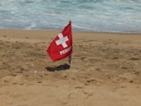 A red warning flag labeled 'PERIGO' with a white cross symbol is planted in the sand on a beach. The ocean waves are visible in the background, approaching the shoreline.