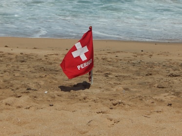 A red warning flag labeled 'PERIGO' with a white cross symbol is planted in the sand on a beach. The ocean waves are visible in the background, approaching the shoreline.