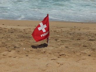 A red warning flag labeled 'PERIGO' with a white cross symbol is planted in the sand on a beach. The ocean waves are visible in the background, approaching the shoreline.