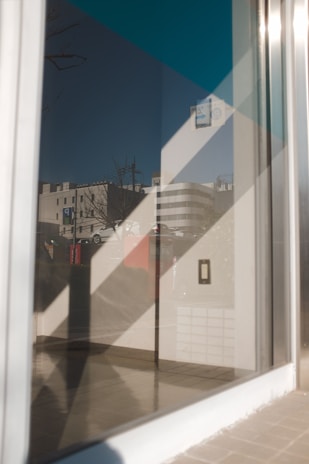 A reflective surface shows the exterior view of a modern urban area. The reflection includes buildings with glass windows, a parked car, and leafless tree branches. Geometric patterns are visible on the glass, creating an artistic overlay.