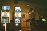 A man is playing darts in a dimly lit room. There are three illuminated electronic dartboards on the wall in front of him, displaying scores and game information. The man is standing to the right, poised to throw a dart. Various posters and a sports jersey are visible on the walls.